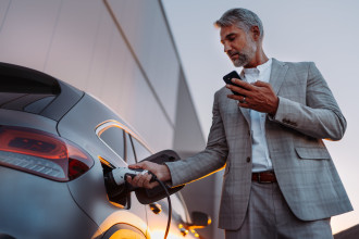 A man who plugs in his electric car. He wears a grey suit and holds a mobile phone in his other hand.