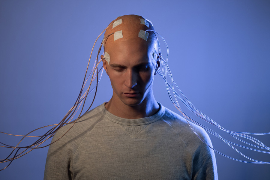 A person with many sensors on his head, from which cables run to the edge of the picture. The background is blue