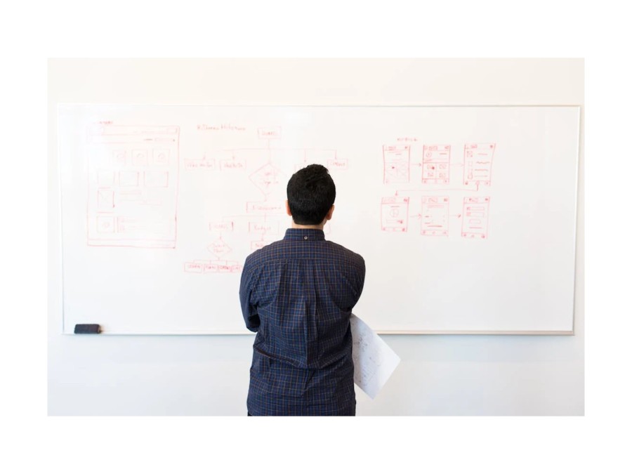 Image of a person standing in front of a white board with red drawing, while they hold a piece of paper.