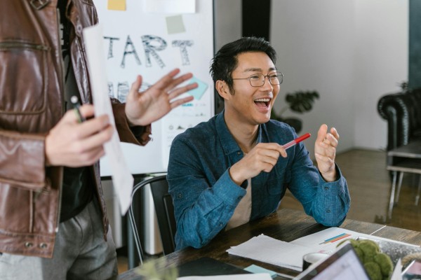 Image of two people in a meeting room, where one person is sitting, smiling and holding a red pen and another person standing next to him with a brown jacket.