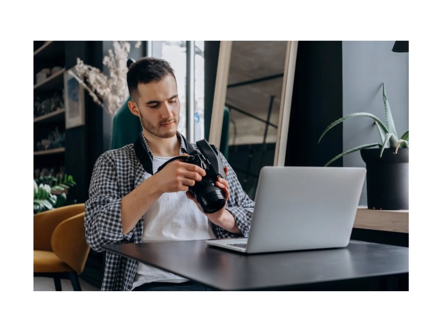 Picture of a Man sitting infront of a Laptop, that sits on a Table, holding a Camera