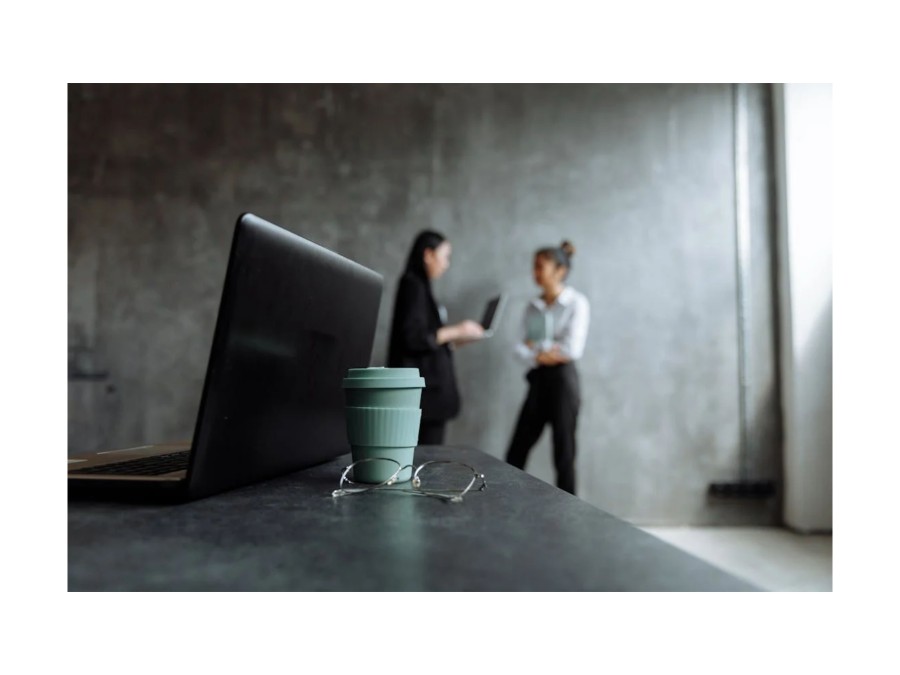 A laptop with a cup of coffe and glasses in focus, while two women are in the background and unfocussed