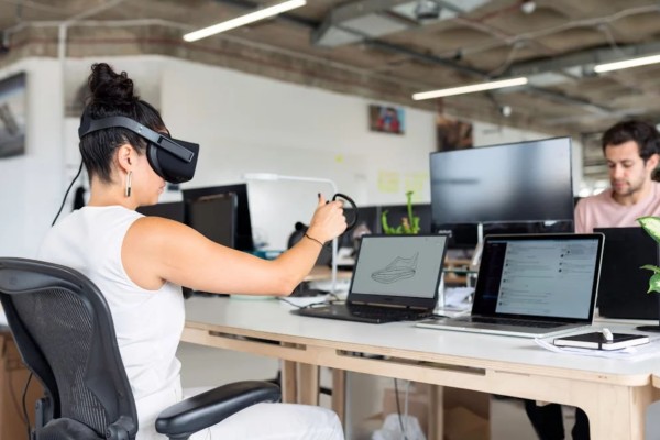 Photo of a Woman in an Office With A Man Wearing a VR Headset Sitting Infront of her Laptop Depicting  Shoe on the Display.